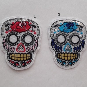 Peut inclure: Deux patchs brodés de crânes de sucre, l'un avec une fleur rouge et l'autre avec une fleur bleue. Les deux patchs sont blancs avec des accents de paillettes argentées.