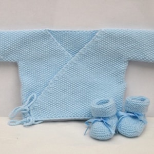 Op de afbeelding: Een lichtblauwe gebreide babytrui met een wikkelgesp en bijpassende slofjes.