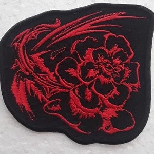 Puede incluir: Parche de tela negro con un diseño de flor roja bordada. La flor es estilizada y tiene un diseño de remolino.