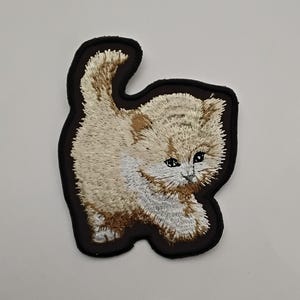 Peut inclure: Un patch brodé représentant un chaton blanc et brun avec une bordure noire. Le chaton est tourné vers la droite de l'image et sa queue est enroulée derrière son dos.