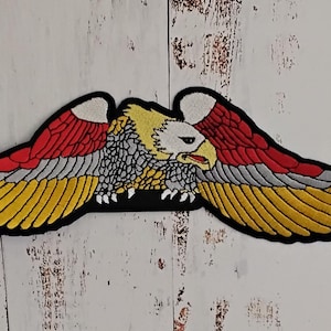Könnte beinhalten: Ein gestickter Adler-Patch mit ausgebreiteten Flügeln in den Farben Rot, Weiß und Gold. Der Adler hat einen weißen Kopf und einen schwarzen Schnabel.