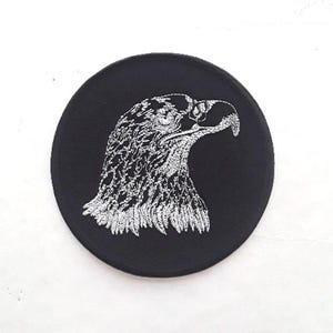 May include: A black embroidered patch with a white eagle head design. The eagle's head is facing right and has a detailed design.