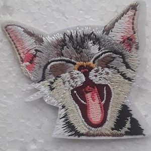 Può includere: Toppa ricamata da stirare con un gatto grigio e bianco con la bocca aperta e la lingua di fuori. Il gatto ha il naso rosa e gli occhi neri.