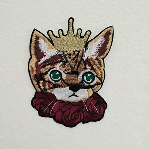 May include: Embroidered patch featuring a cat's face wearing a gold crown and a red rose collar. The cat has green eyes and brown and tan fur. The patch is designed for application on clothing or accessories.