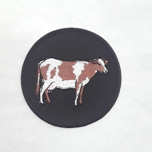 Peut inclure: Un patch circulaire noir brodé d'une vache brune et blanche avec des contours noirs.