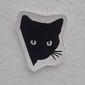 Può includere: Una toppa da stirare ricamata in bianco e nero con il viso di un gatto. Il gatto ha occhi bianchi e un baffo bianco.