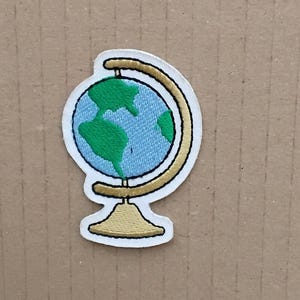 Pode incluir: Um remendo bordado branco de um globo em um suporte. O globo é azul e verde com um suporte dourado.