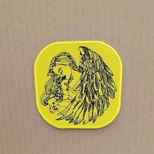 Op de afbeelding: Gele stoffen patch met een zwarte lijntekening van een engel met vleugels die een klein kind vasthoudt. De engel heeft lang haar en kijkt naar het kind.