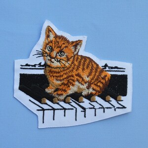 May include: Embroidered patch featuring a ginger tabby kitten sitting on a piano keyboard. The kitten has detailed fur and is set against a white background with black and tan piano keys. The patch is likely for clothing or accessories.