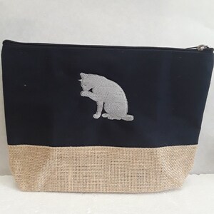 May include: A black and tan zippered pouch with a gray embroidered cat design. The pouch is made of canvas and burlap.