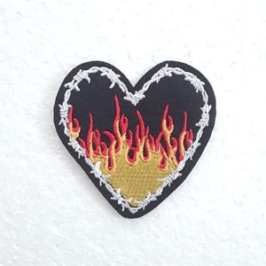 Puede incluir: Parche negro en forma de corazón con un borde de alambre de púas y llamas rojas y amarillas bordadas en el interior.