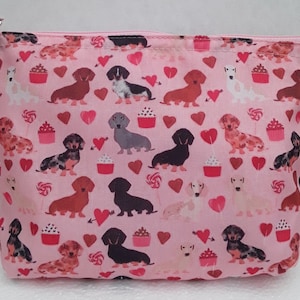 Könnte beinhalten: Rosa Reißverschluss-Tasche mit einem Dackel-Druck. Der Druck zeigt Dackel in verschiedenen Farben und Posen, mit Herzen, Cupcakes und Lutschern auf rosa Hintergrund.