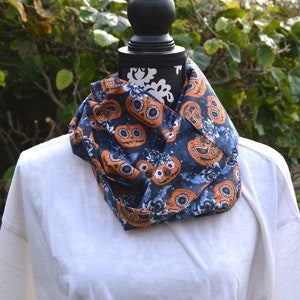 Könnte beinhalten: Ein blauer Infinity-Schal mit einem Muster aus orangefarbenen und weißen Zucker-Schädel-Kürbissen. Der Schal befindet sich auf einer weißen Puppe.
