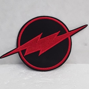 Può includere: Toppa ricamata rossa con un cerchio nero e un fulmine rosso al suo interno. Il fulmine è inclinato in diagonale attraverso il cerchio.