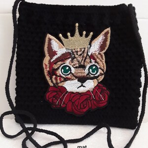 Op de afbeelding: Zwarte gehaakte tas met een kat die een kroon draagt en rode rozen geborduurd op de voorkant. De tas heeft een zwarte band.