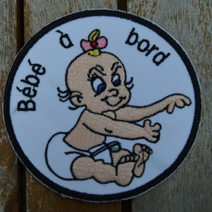 Peut inclure: Un écusson rond blanc avec une bordure noire représentant un bébé fille dessin animé avec un nœud rose dans les cheveux. Le texte "Bébé à bord" est brodé en noir autour du bébé.
