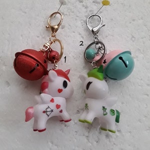 Peut inclure: Deux porte-clés en forme de licorne blanche avec des motifs différents. Le porte-clés de gauche a un motif cœur rouge et flèche et un charme de cloche rouge. Le porte-clés de droite a un motif trèfle vert et pomme et un charme de cloche bleu et rose.