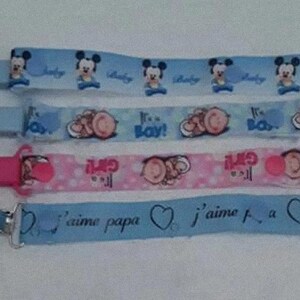 Pode incluir: Quatro prendedores de chupeta azuis e rosa com diferentes designs. O prendedor superior apresenta um design de Mickey Mouse com o texto "It's a Boy!" O segundo prendedor apresenta um design de bolinhas rosa e branco com o texto "It's a Girl!" O terceiro prendedor apresenta um design azul e branco com o texto "J'aime Papa" e um coração. O quarto prendedor apresenta um design azul e branco com o texto "J'aime La" e um coração.
