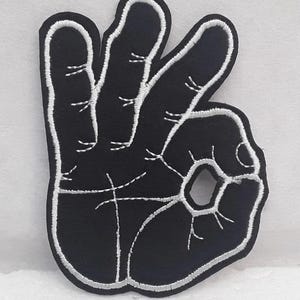 Op de afbeelding: Zwarte geborduurde strijkpatch van een hand die het 'OK'-teken maakt. De hand is wit omrand.