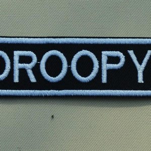 Peut inclure: Un écusson brodé noir et blanc avec le mot "DROOPY" en lettres blanches.
