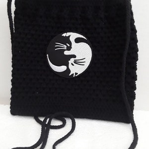 Könnte beinhalten: Schwarze gehäkelte Tasche mit einem weißen Yin-Yang-Symbol, das zwei sich gegenüberstehende Katzen zeigt. Die Tasche hat einen langen schwarzen Riemen.