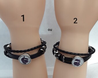 Può includere: Due braccialetti in pelle nera con perline argentate e un ciondolo argentato con la silhouette di una testa umana. I braccialetti sono mostrati su due braccia di manichino. Il testo "1" e "2" è scritto sulle braccia, e la parola "ou" è scritta tra le braccia.