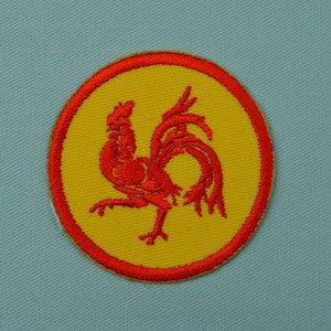 Peut inclure: Un coq rouge brodé sur un cercle jaune avec une bordure rouge. Le coq est tourné vers la droite et a les plumes de sa queue déployées.