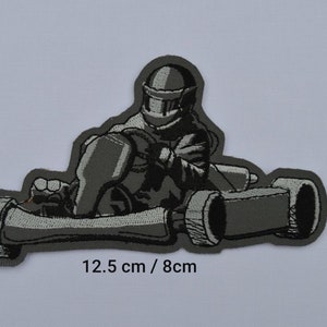 Könnte beinhalten: Ein grauer gestickter Aufnäher mit einer Person, die einen Helm trägt und einen Go-Kart fährt. Der Aufnäher misst 12,5 cm x 8 cm.
