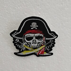 Peut inclure: Patch brodé noir et blanc d'un crâne de pirate portant un chapeau noir avec un bandana rouge et des os croisés. Le crâne tient deux épées croisées devant lui.