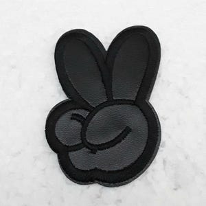 Peut inclure: Patch en cuir noir avec un signe de paix en forme de main avec un œil qui cligne.