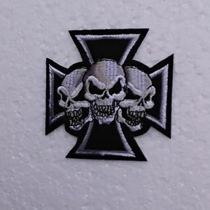 Pode incluir: Um remendo bordado em preto e branco com três caveiras em um design de cruz. As caveiras são brancas com olhos e dentes pretos. A cruz é preta com costuras brancas.