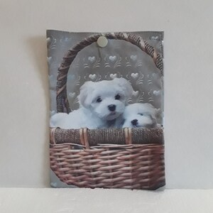 May include: A gray fabric pouch with a white button closure. The pouch features a printed image of two white puppies in a brown wicker basket with a gray and white heart pattern background.