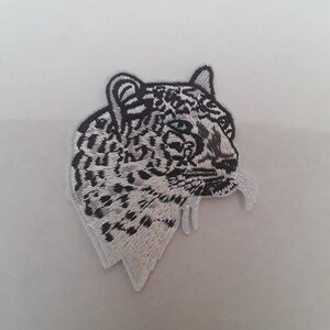 Peut inclure: Patch brodé blanc représentant la tête d'un léopard des neiges avec des marques noires et un œil bleu.