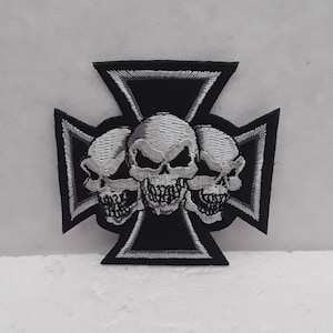 Könnte beinhalten: Schwarz-weiß gestickter Patch mit drei Schädeln in Silber, zentriert auf einem schwarzen Eisenkreuz mit silbernen Verzierungen.