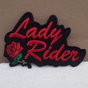 Op de afbeelding: Rode geborduurde patch met de tekst "Lady Rider" in een cursief schrift. Een rode roos met groene bladeren bevindt zich links van de tekst. De patch is op een zwarte achtergrond.