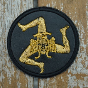 May include: A black leather patch with a gold embroidered three-legged figure, known as the Trinacria, a symbol of Sicily.