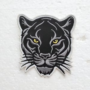 Peut inclure: Écusson brodé représentant une tête de panthère. L'écusson a un fond noir avec des détails gris et une bordure grise. Les yeux de la panthère sont jaunes. Ce patch peut être repassé ou cousu sur des vêtements ou des accessoires.