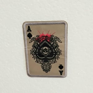 Può includere: Una toppa ricamata beige con un designo di carta da gioco nera asso di picche. La carta presenta un teschio e ossa incrociate al centro, circondati da una ghirlanda di fiori e due aquile con le ali spiegate. La carta è bordata da un contorno bianco.