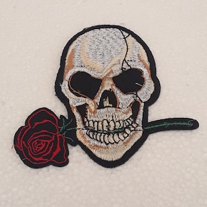 Può includere: Toppa ricamata con un teschio bianco con un'orbita nera e una rosa rossa in bocca. Il teschio sorride e la rosa è di colore rosso scuro.