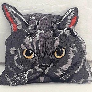 Op de afbeelding: Gestikt strijkbaar patch van een grijze kat met gele ogen en rode oren. De kat kijkt recht naar de kijker.