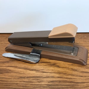 May include: A brown metal stapler with a beige plastic top and a silver staple remover. The stapler is on a wooden surface.