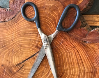 May include: A pair of black handled scissors with silver blades on a wooden surface with concentric rings. The scissors are open and the blades are pointing towards the bottom of the image.