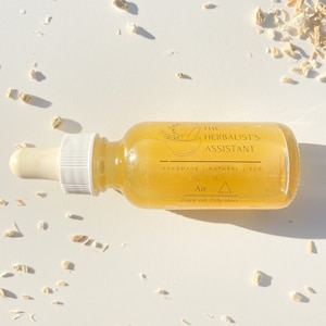 Op de afbeelding: Een gele glazen fles met een druppelaar en een etiket met de tekst "The Herbalist's Assistant" en "Air Face oil - Oily skin". De fles staat op een wit oppervlak met kleine stukjes gedroogde kruiden eromheen.