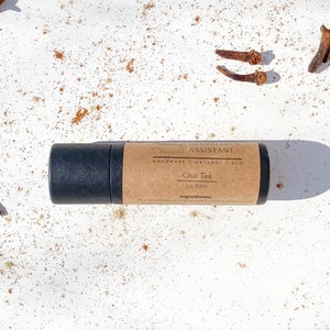 Könnte beinhalten: Eine schwarze Lippenbalsam-Tube mit einem braunen Etikett mit der Aufschrift "HERBALIST ASSISTANT Chai Tea Lip Balm". Der Balsam ist von verstreuten Gewürzen und ganzen Nelken auf einer weißen Oberfläche umgeben. Die Verpackung ist umweltfreundlich.