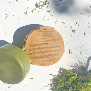 May include: A brown paper-wrapped bar of soap with a green label that reads "The Herbalist's Assistant Handmade Natural Soap Earth Conditioner Bar" and lists the ingredients. The soap is sitting on a white surface with green leaves scattered around it.