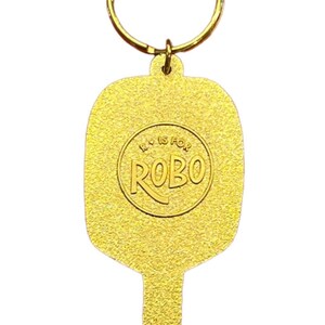 K&ouml;nnte beinhalten: Goldfarbener Schl&uuml;sselanh&auml;nger aus Metall in Form eines Paddels mit einem runden Logo, auf dem "R is for ROBO" steht.