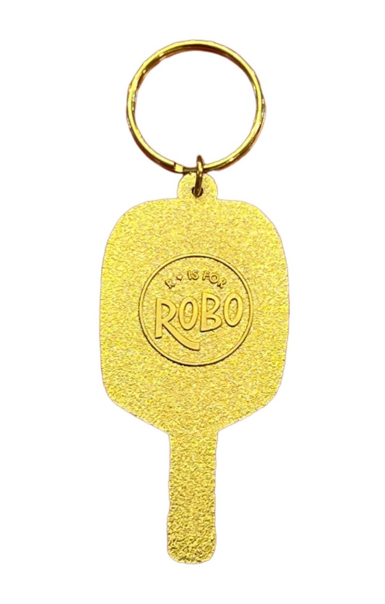 K&ouml;nnte beinhalten: Goldfarbener Schl&uuml;sselanh&auml;nger aus Metall in Form eines Paddels mit einem runden Logo, auf dem "R is for ROBO" steht.