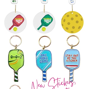 K&ouml;nnte beinhalten: Ein Set an Pickleball-Schl&uuml;sselanh&auml;ngern, Ornamenten und Aufklebern. Die Schl&uuml;sselanh&auml;nger zeigen einen Pickleball-Schl&auml;ger mit dem Text "0-0-2", "One More Game" und "Stay Out of the Kitchen". Es gibt auch einen gelben Pickleball-Schl&uuml;sselanh&auml;nger. Die Ornamente zeigen einen Pickleball-Schl&auml;ger mit dem Text "Pickleballin' Around the Christmas Tree". Die Aufkleber zeigen einen Pickleball-Schl&auml;ger mit dem Text "One More Game", "Stay Out of the Kitchen" und "0-0-2".