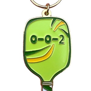 K&ouml;nnte beinhalten: Ein gr&uuml;ner und goldener Pickleball-Schl&uuml;sselanh&auml;nger mit dem Text "0-0-2" auf dem Schl&auml;ger. Der Schl&uuml;sselanh&auml;nger hat einen goldenen Metallring.