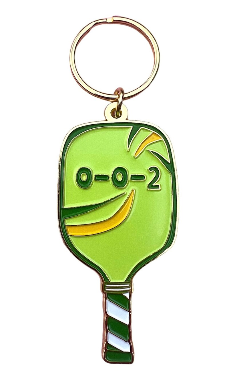 K&ouml;nnte beinhalten: Ein gr&uuml;ner und goldener Pickleball-Schl&uuml;sselanh&auml;nger mit dem Text "0-0-2" auf dem Schl&auml;ger. Der Schl&uuml;sselanh&auml;nger hat einen goldenen Metallring.